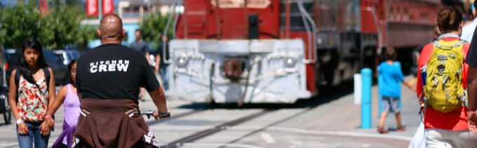 A man riding a bike in a sidepath next to a set of railroad tracks is visible near a train parked nearby. Several people walking nearby are also visible.