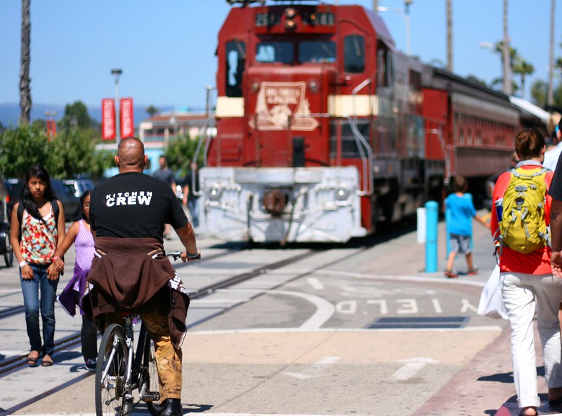 A man riding a bike in a sidepath next to a set of railroad tracks is visible near a train parked nearby. Several people walking nearby are also visible.